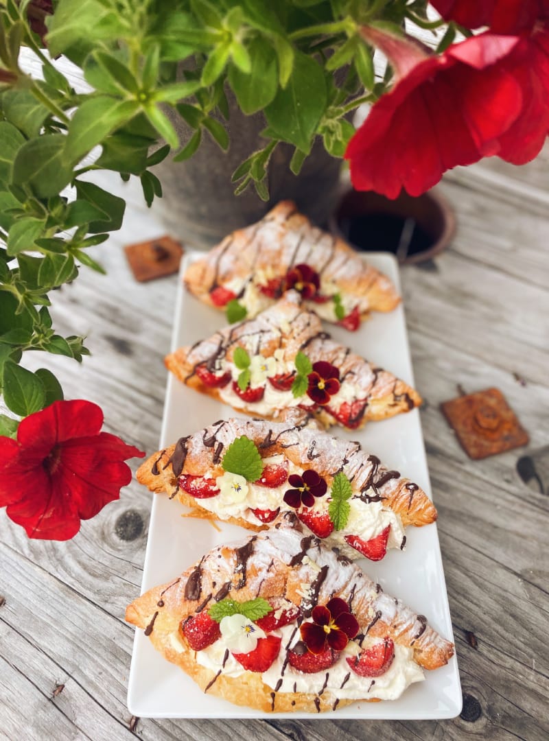 Nygräddade croissanter med grädde, jordgubbar, choklad och ätbara blommor på ett fat utomhus.
